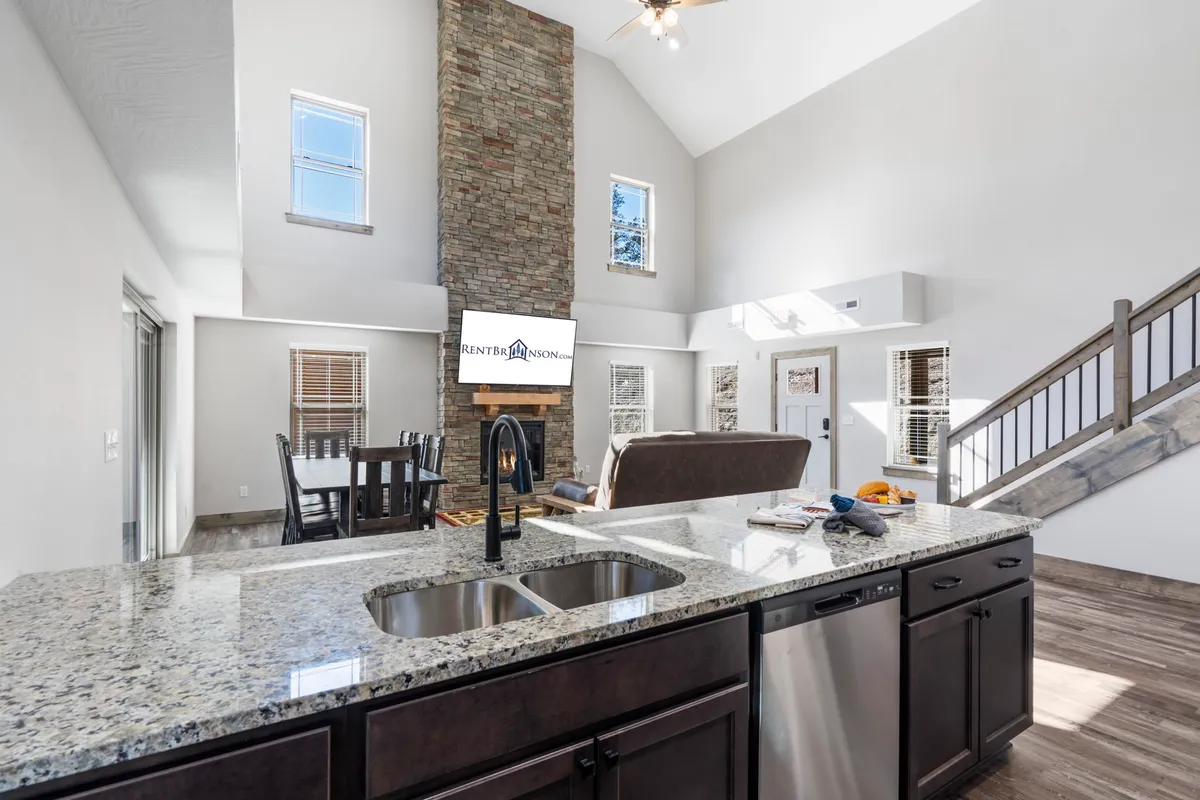A spacious, open-concept interior showcasing a stone fireplace, a kitchen island with bar seating, a dining area, and large windows that welcome natural light. The arrangement emphasizes togetherness with the kitchen, dining, and living spaces flowing seamlessly into each other, highlighted by cozy furnishings and modern touches.