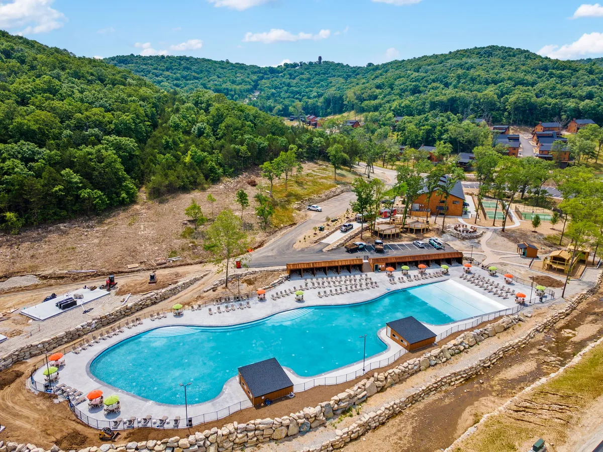 A large outdoor swimming pool with surrounding green hills and a nearby deck area furnished with lounge chairs and wooden structures, set against a backdrop of beautiful natural scenery.