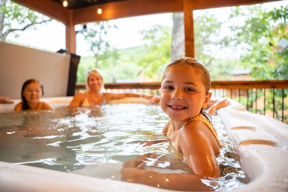 A family scene unfolds in a hot tub set amidst natural surroundings. A smiling child and two women relax together, capturing moments of laughter, bonding, and relaxation under the open sky.
