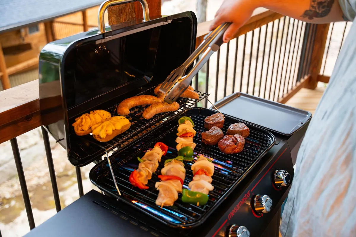 A lively outdoor barbecue scene with a grill loaded with skewers, sausages, and vegetables. People gather around, enjoying food and conversation as twilight approaches. The backdrop of natural greenery suggests a relaxed, community-oriented atmosphere.