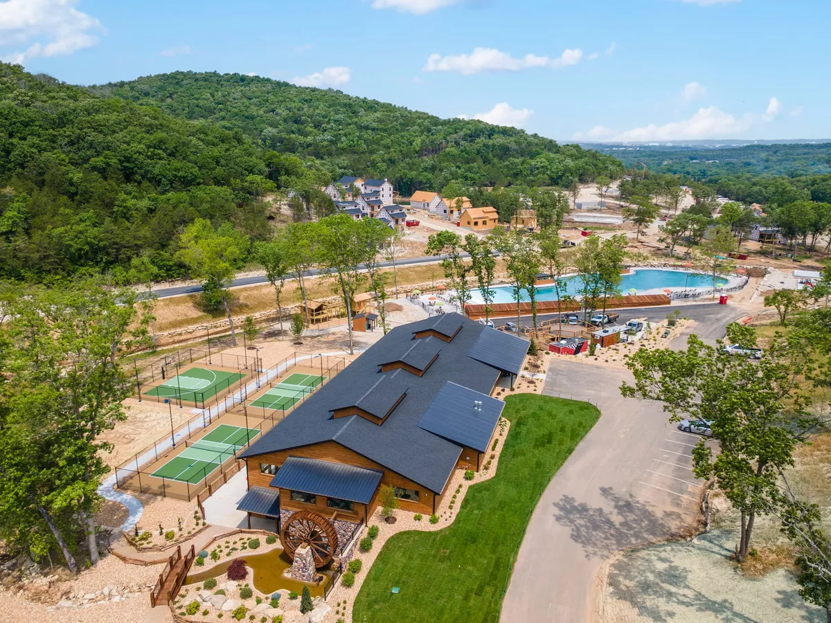 The vibrant community center stands at the heart of Tall Timbers Camp, with a sparkling swimming pool, tennis courts, and winding nature trails visible. People can be seen enjoying the various amenities in a lush, green setting, highlighting the resort-style activities available to guests.