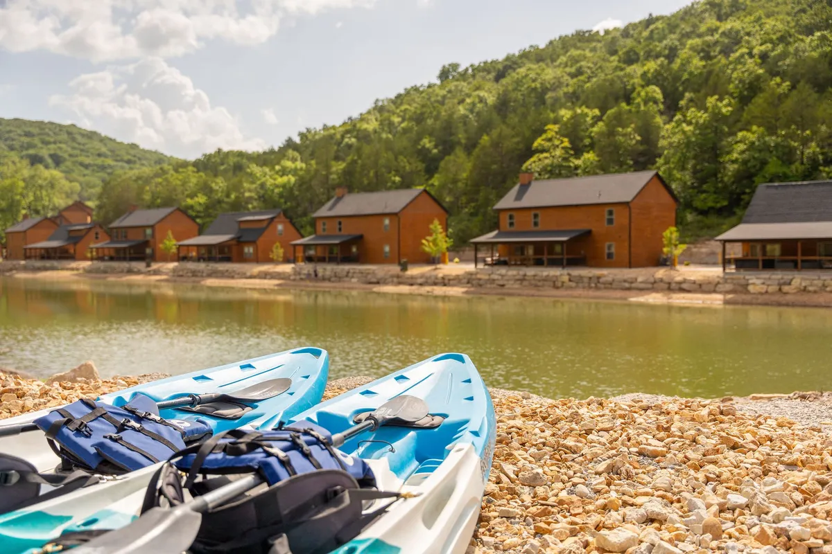 This image features kayaks resting near rustic cabins on the shores of a serene lake, set against a mountain backdrop and surrounded by lush greenery. The scene evokes adventure and relaxation, highlighting the area's natural beauty and water-based activities.