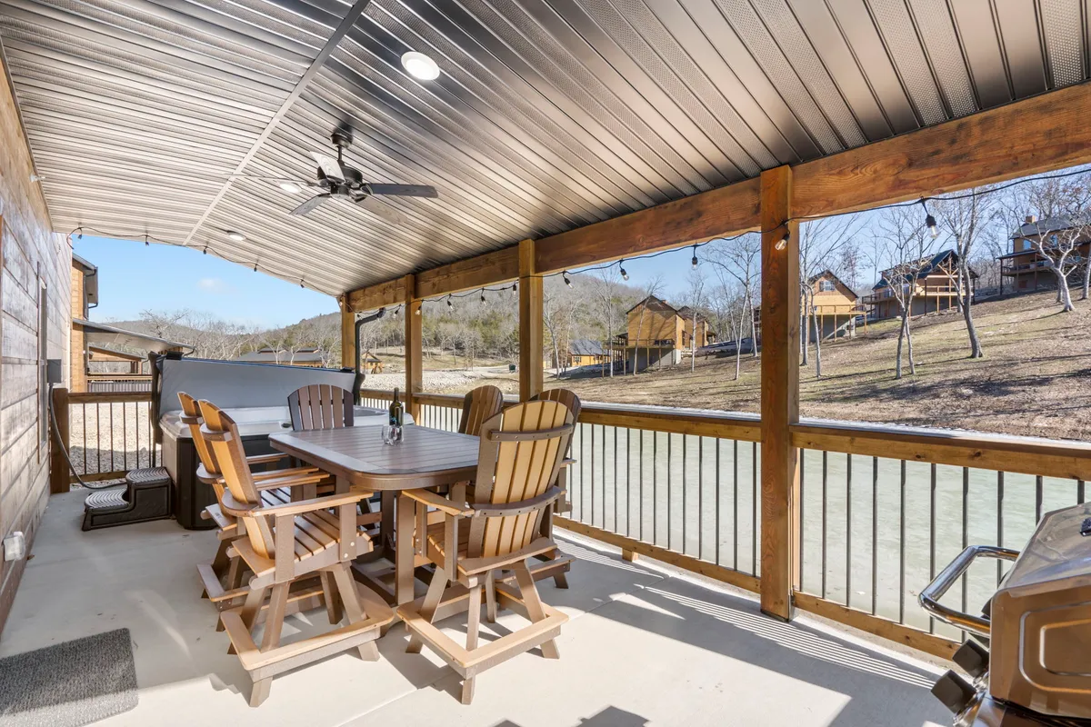 An inviting outdoor dining area on a wooden deck, complete with a dining table, wooden chairs, and a nearby grill looking out over a scenic river view. The space is set up for lively barbecues and group gatherings.