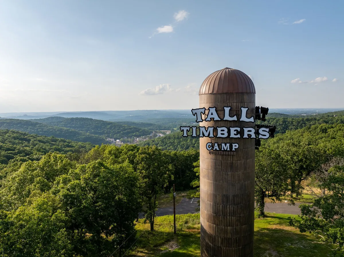 A wide view of the Tall Timbers Camp water tower rising above the lush forest, with distant hills in the background. The expansive sky hints at sunset, and the landscape invites exploration of the surrounding woods and trails.